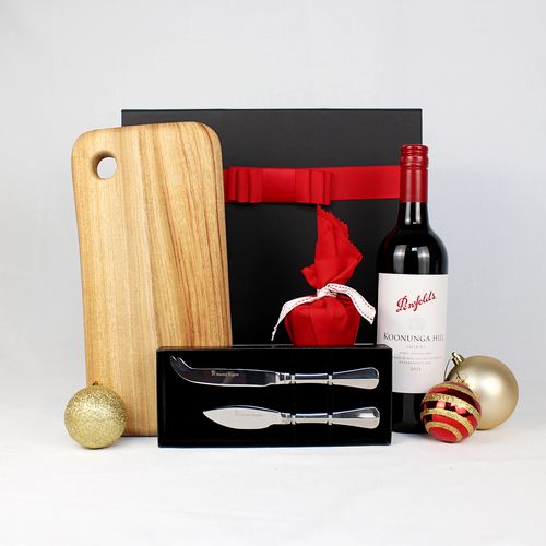 Black gift box with red ribbon behind a bottle of Australian wine, handmade timber cheeseboard, stainless steel cheese knife set and a small Christmas pudding in red fabric