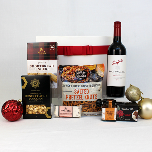 white and red gift box standing behind a range of festive treats including a small Christmas pudding and sauce, pretzels, honey coated popcorn, shortbread and nougat alongside a bottle of Australian wine surrounded by Christmas baubles.