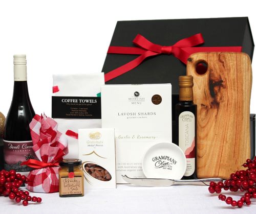a black gift box with red ribbon standing behind a bottle of Australian red wine, a handmade timber cheese board with stainless steel cheese knife, infused olive oil with white dipping bowl, lavosh shards, smoked almonds, linen hand towel and Christmas pudding in red fabric with sauce
