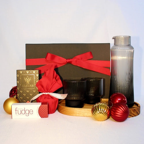 black gift box with red festive ribbon standing behind a handmade timber tray with ribbed glass carafe and glasses pair sitting upon it alongside an Australian made Christmas pudding, chocolate bar and fudge surrounded by Christmas baubles