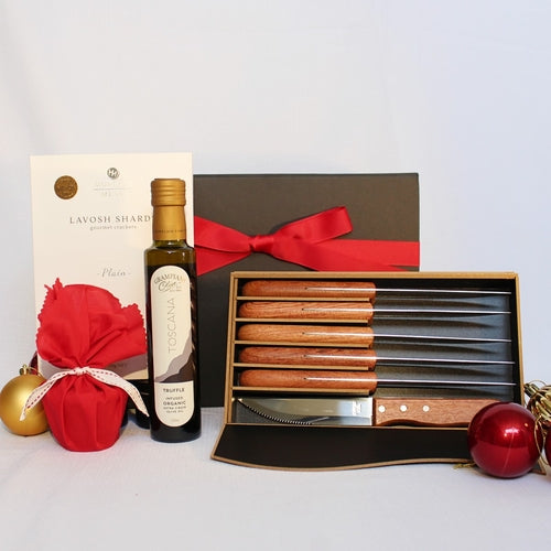 Black gift box with red ribbon behind a set of 6 timber and steel steak knives alongside a bottle of infused olive oil, a box of lavish shards and a Christmas pudding