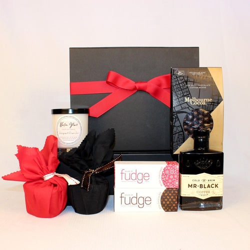 Black gift box with festive red ribbon standing behind a bottle of Coffee Liqour and a range of sweets - two Christmas puddings, two bars of fudge, a bag of chocolate coated coffee beans and a luxury hand poured candle.