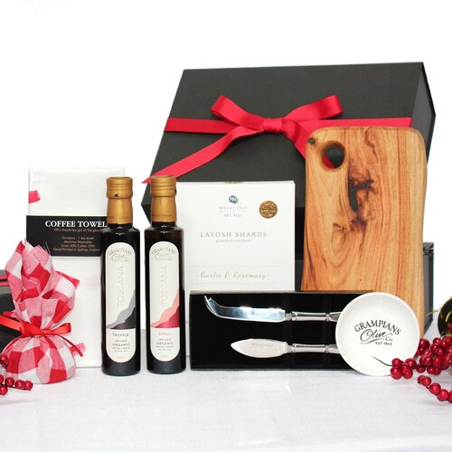 a black gift box with a red ribbon behind a handmade timber platter accompanied by a cheese knife set, infused olive oil bottles x2, dipping bowl, box of lavosh shards, handpainted linen hand towel and a Christmas pudding in red fabric