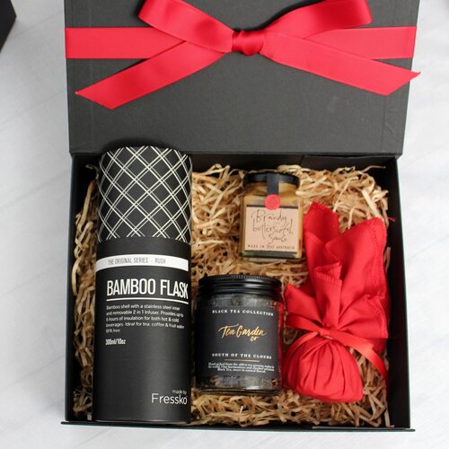 black gift box with festive red ribbon opened to show contents of a reusable, sustainable bamboo drink flask accompanied by Christmas pudding in red fabric, pudding sauce and a jar of loose leaf tea