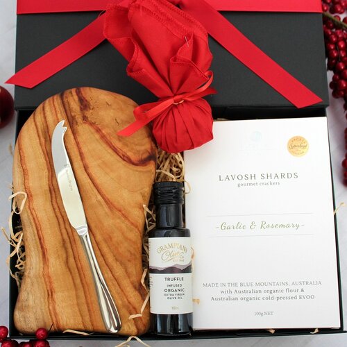 An opened black gift box with red ribbon showing contents of a timber cheese board, stainless steel cheese knife, infused olive oil, box of lavosh shards and a small Christmas pudding in red fabric