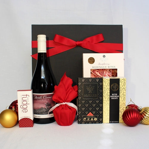 black gift box with red ribbon surrounded by christmas baubles and products - Australian red wine, christmas pudding, chocolate, fudgr, shortbread and raspberry meringue bites.