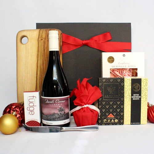 black gift box with red ribbon standing behind a range of Christmas products. Handmade timber serving board with stainless steel knife, meringue bites, chocolate, fudge, shortbread and Australian wine