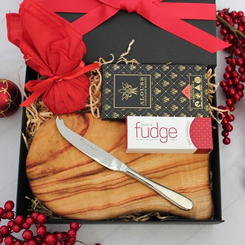 black gift box with red ribbon opened showing contents within - a handmade timber cheese board with stainless steel cheese knife on the board Australian chocolate, fudge and Christmas pudding in red fabric with red holly surrounding the set up