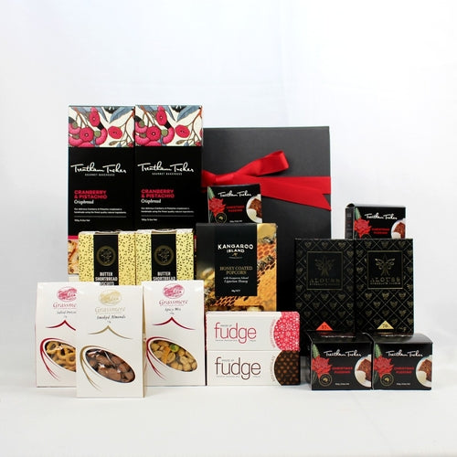 black gift box with red ribbon standing behind a range of food hamper products - boxes of crispbread, shortbread, honey coated popcorn, pretzels, spicy mix, smoked almonds, chocolate and Christmas Puddings with black, red and gold packaging