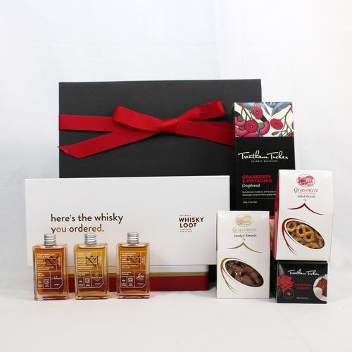 black gift box with red ribbon standing behind a box of crispbread in black and red box with small Christmas pudding, pretzels and smoked almonds alongside a box of 3 whisky sample bottles